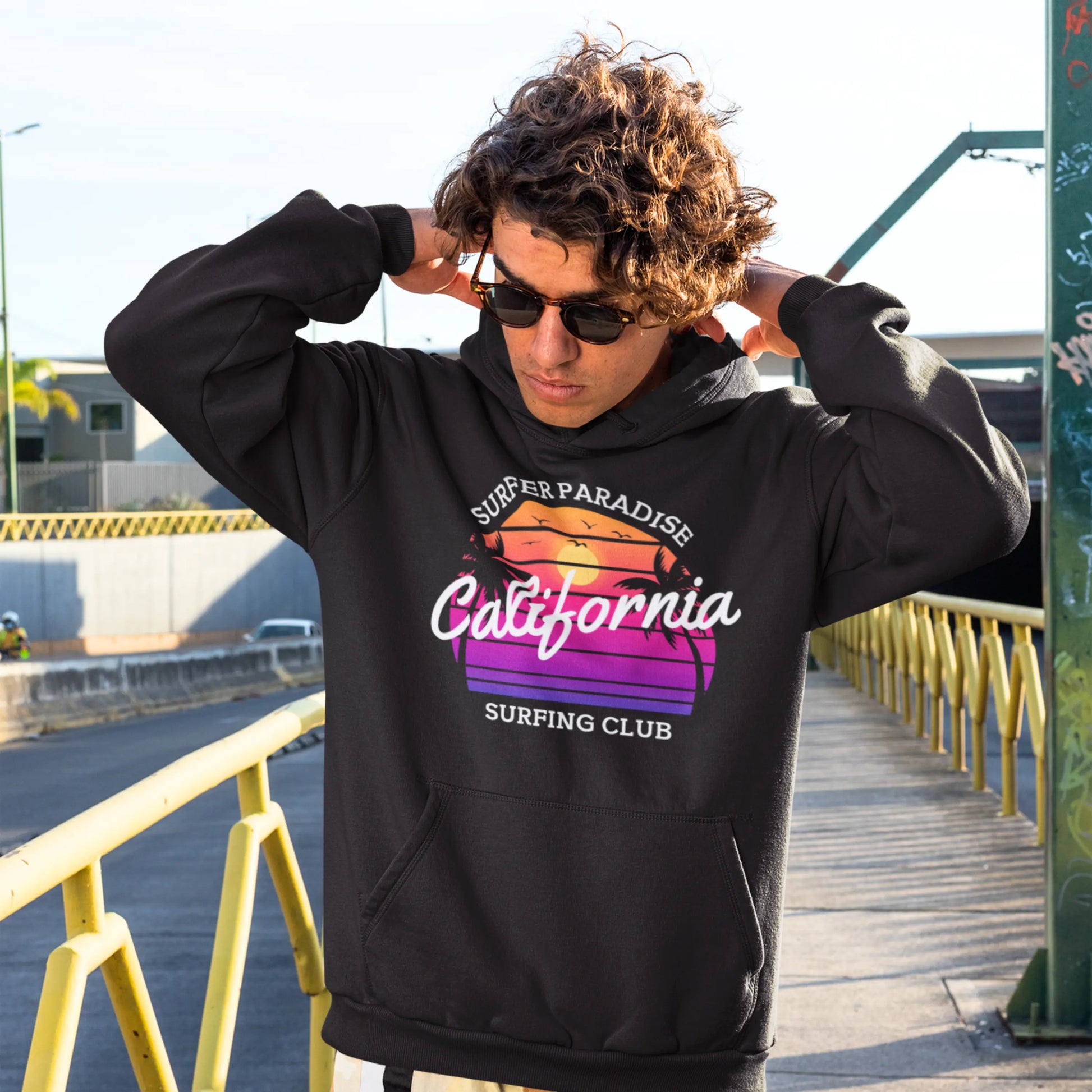 A young man with curly hair stands on a sunlit city bridge, casually adjusting his black bucket hat while wearing dark sunglasses. He's dressed in a black hoodie with a vibrant graphic on the front that reads 'Surfer Paradise California Surfing Club' in bold, gradient colors of pink, orange, and purple, evoking a sense of urban coolness and casual street style.