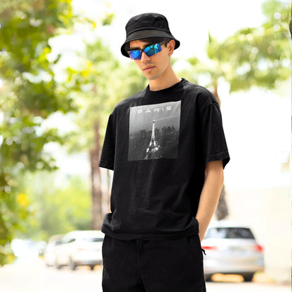 A young man stands casually outdoors, wearing a black bucket hat and sunglasses with blue reflective lenses. He is dressed in a black t-shirt with a square white graphic that has the word 'PARIS' and an image of the Eiffel Tower, symbolizing a classic, urban style.