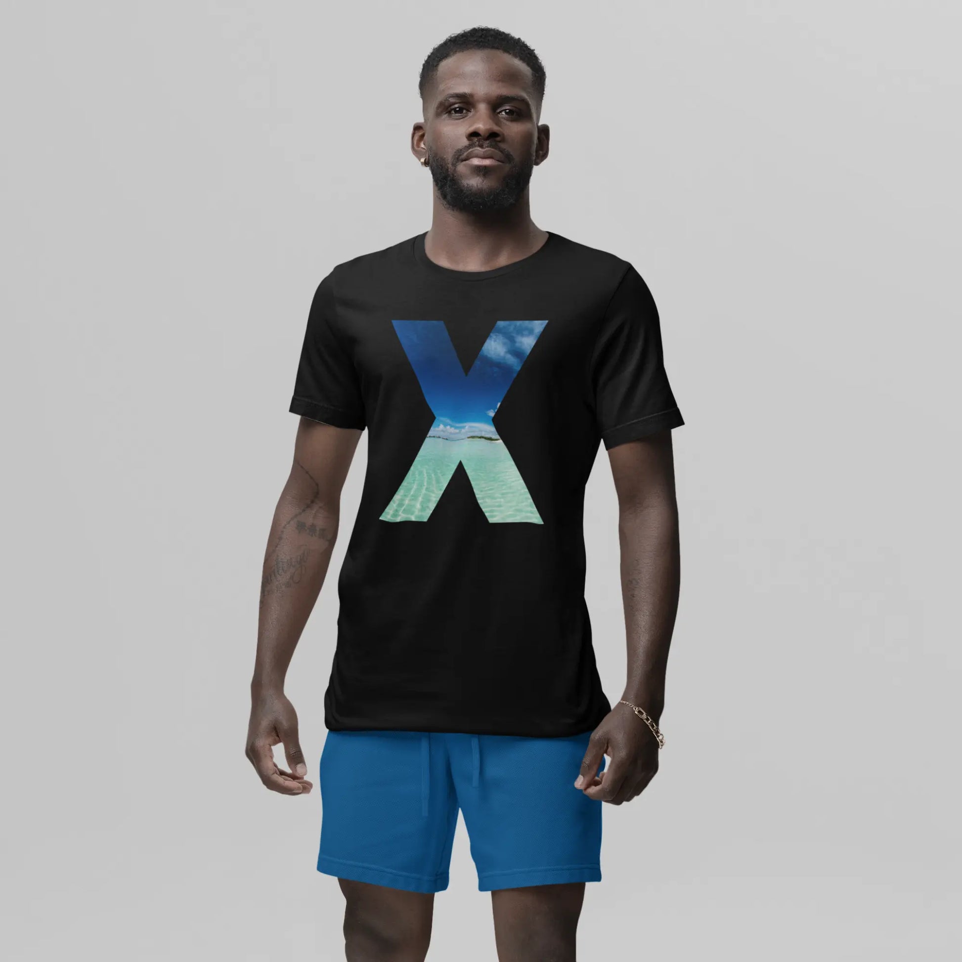  A confident man stands outdoors in a relaxed stance, wearing a black t-shirt featuring a stylized 'X' with a sea wave print within it. 