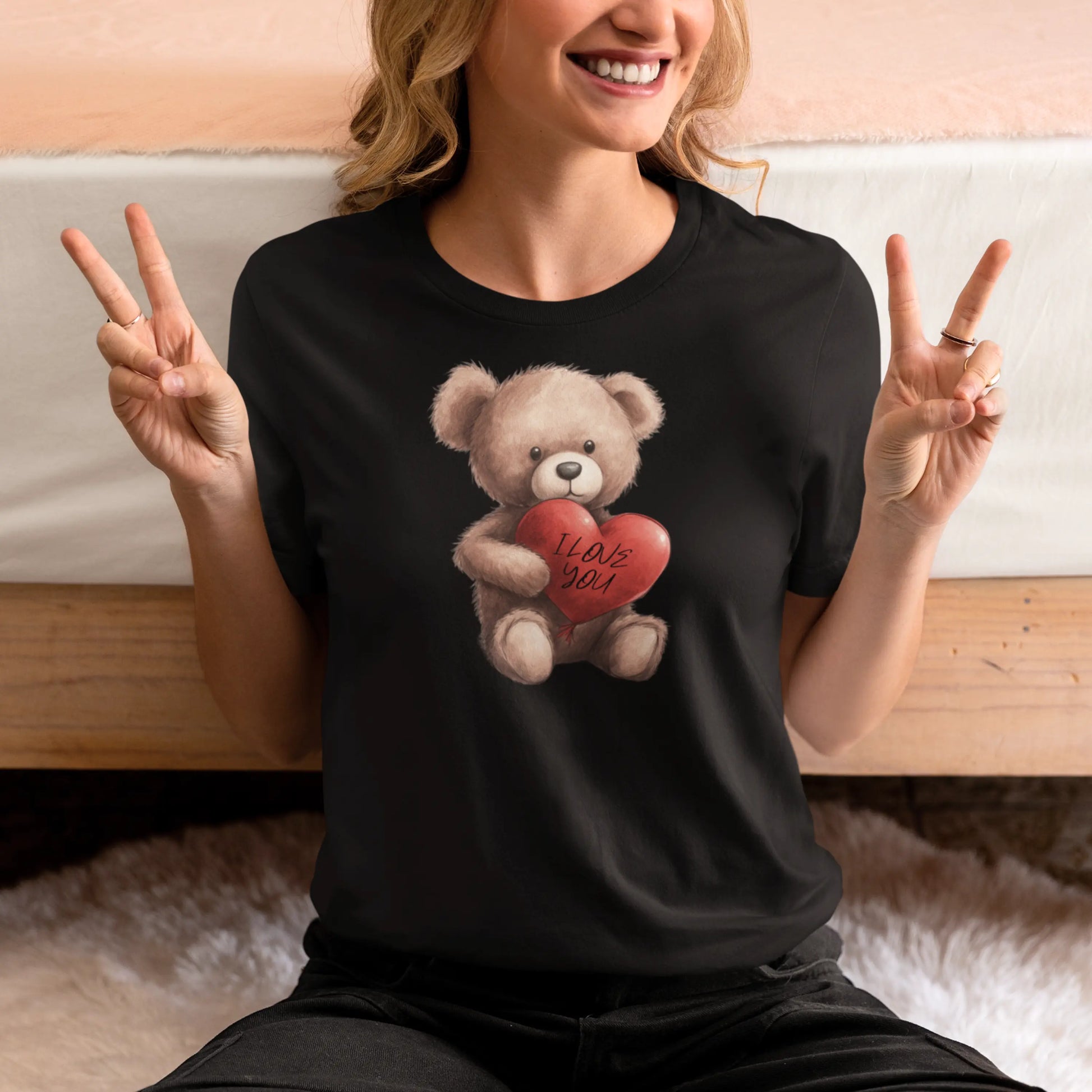 A woman in a black t-shirt is making a peace sign with both hands. The t-shirt features a cute, soft-focus graphic of a teddy bear holding a heart that says 'I Love You.' She is seated with a warm, genuine smile.