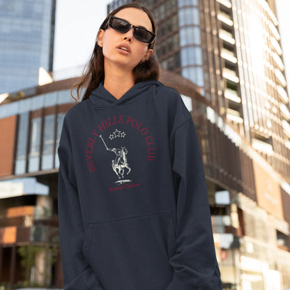A confident woman stands in an urban environment wearing a dark navy blue hoodie with 'Beverly Hills Polo Club' and 'Limited Edition' in red text around a polo player on horseback emblem. She is accessorized with sleek sunglasses, and the city's tall buildings provide a modern backdrop to her street-style outfit.