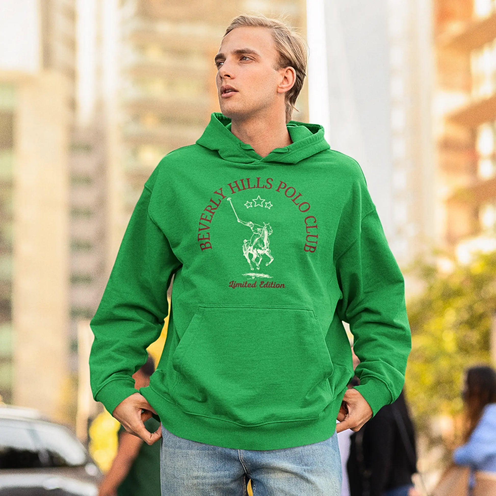 A man with slicked-back blonde hair, gazing thoughtfully into the distance, is wearing a bright green hoodie with 'Beverly Hills Polo Club' and an emblem of a polo player on horseback embroidered on the front, labeled 'Limited Edition'. The vibrant green of the hoodie stands out against the urban backdrop of towering buildings and a busy street.