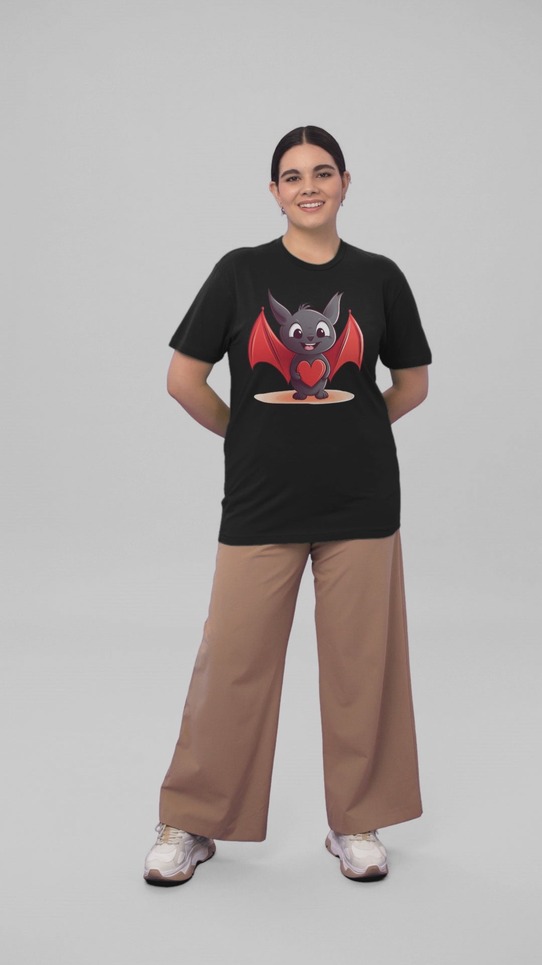 A smiling woman in a studio psing with a black t-shirt adorned with a cute cartoon bat character in the center holding a red heart, showcasing a playful and loving design.
