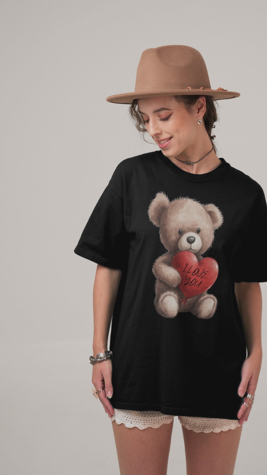 A woman wearing a black oversized t-shirt with a graphic of a cuddly teddy bear holding a red heart that says 'I Love You', paired with a delicate lace skirt and layered bracelets, creating a blend of soft and edgy styles.